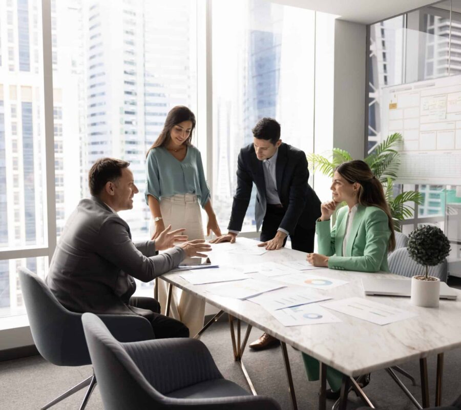 Multiethnic startup developers team gather around boardroom table brainstorming insights Multiethnic company managers startup developers team with middle aged leader boss gather around boardroom table brainstorming insights creating plans reviewing project materials in modern urban office